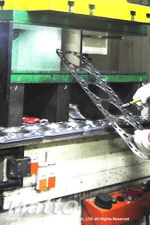 A worker in white gloves operates a machine, holding a large, intricately cut metal piece, likely a gasket or frame, in an industrial setting with heavy machinery in the background.