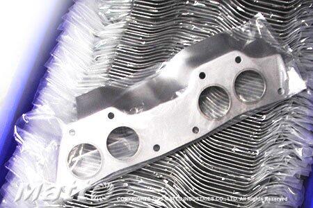 A new, multi-layered metal exhaust manifold gasket with four distinct port openings, lying on a stack of similar gaskets, partially covered by clear plastic packaging, highlighting its metallic construction and precision design.