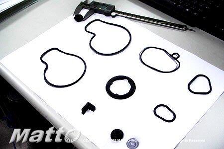 A white sheet of paper displays a set of black rubber O-rings and seals, likely part of a hydraulic pump seal kit, with a metal caliper resting at the top.