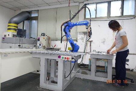 A blue Yaskawa industrial robot, likely engaged in a task like welding, stands positioned over a metal workbench, while a person on the right side of the image interacts with materials on the table, indicating a manufacturing or research setting.
