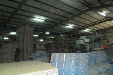 A wide-angle view inside a large industrial warehouse or factory building, revealing a high ceiling with exposed metal trusses and fluorescent lighting. 