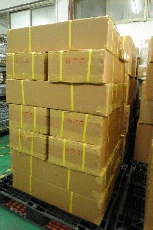A tall stack of cardboard boxes, secured with yellow packing tape, rests on a black plastic pallet in what appears to be a warehouse or storage facility, with other stacked boxes visible in the background.