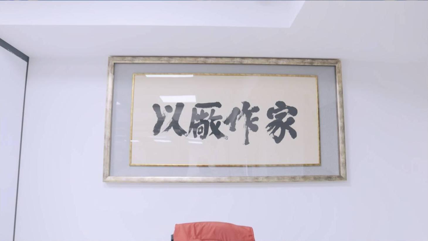 A framed piece of Chinese calligraphy, featuring bold black characters on a light background, hanging on a white wall above a glimpse of a red chair. The text reads "以厰作家" (yǐ chǎng zuò jiā).