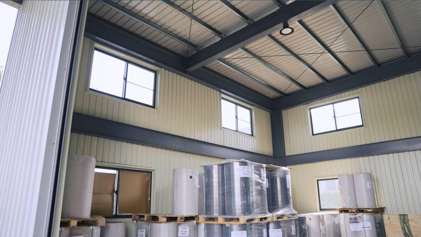 Interior view of a warehouse or industrial building, showcasing a high ceiling with exposed metal beams and roof structure, multiple windows, and large rolls of construction materials, possibly insulation, stacked on wooden pallets across the floor.
