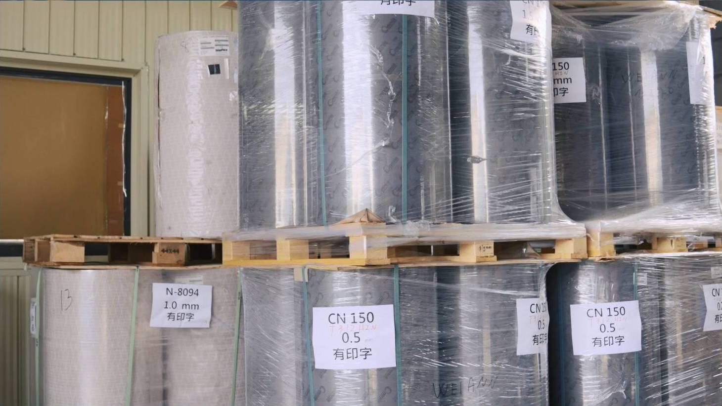 Large rolls of material, possibly self-adhesive paper or labels, are neatly stacked on wooden pallets in what appears to be a warehouse or storage area. The rolls are individually wrapped in clear plastic, and some have labels with text like "N-8094," "CN 150," and measurements such as "1.0 mm" and "0.5."