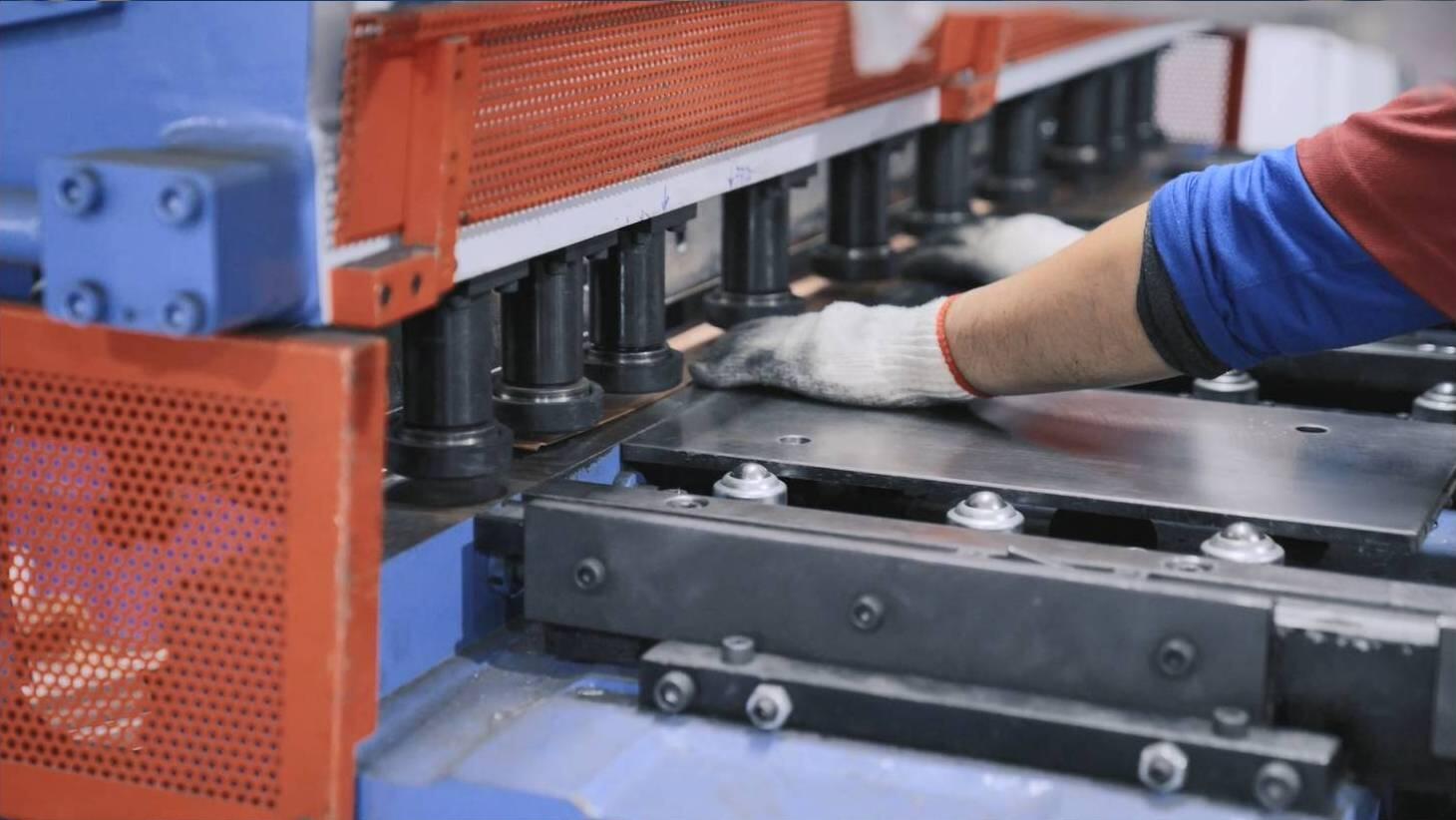 A worker in gloves carefully guides a metal sheet under the bending tools of a large, red and blue industrial press brake machine, preparing it for a precise bend in a manufacturing setting.