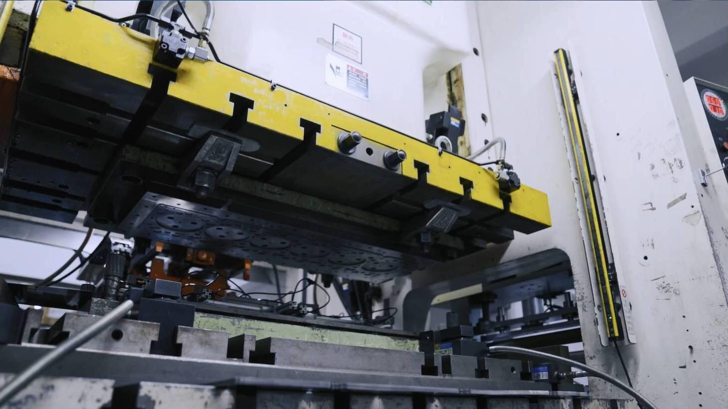 Close-up view of a large yellow and gray industrial metal stamping press with visible dies and automation components, suggesting manufacturing or fabrication.