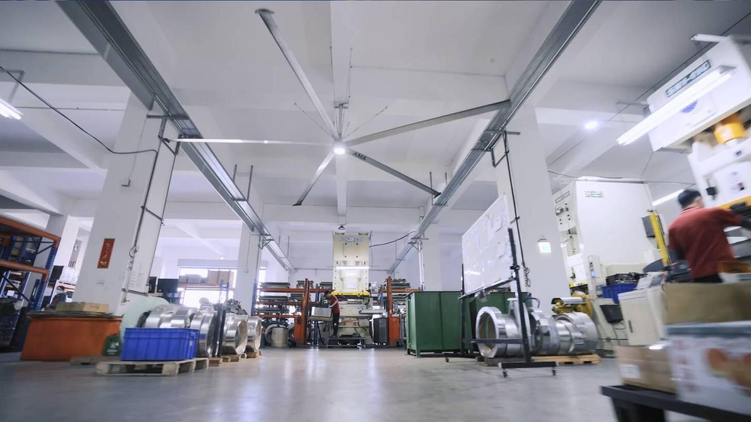  A large industrial facility features a prominent HVLS (High-Volume Low-Speed) ceiling fan for air circulation, with various machinery, stacked materials like metal coils, and a worker in the background, suggesting a manufacturing or warehouse environment.