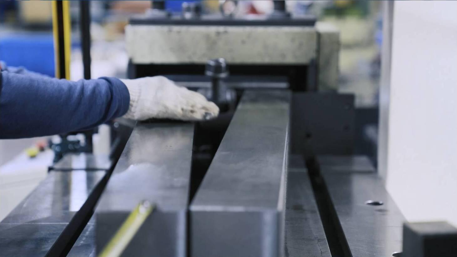 A gloved hand positions a piece of metal on a press brake machine, preparing it for bending, with the machine's upper die visible above.