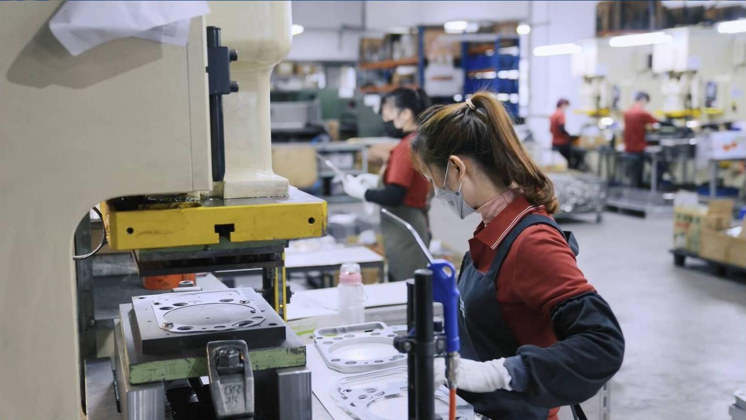  A factory worker, wearing a mask and gloves, operates machinery to manufacture gaskets in a factory setting. Other workers are visible in the background.