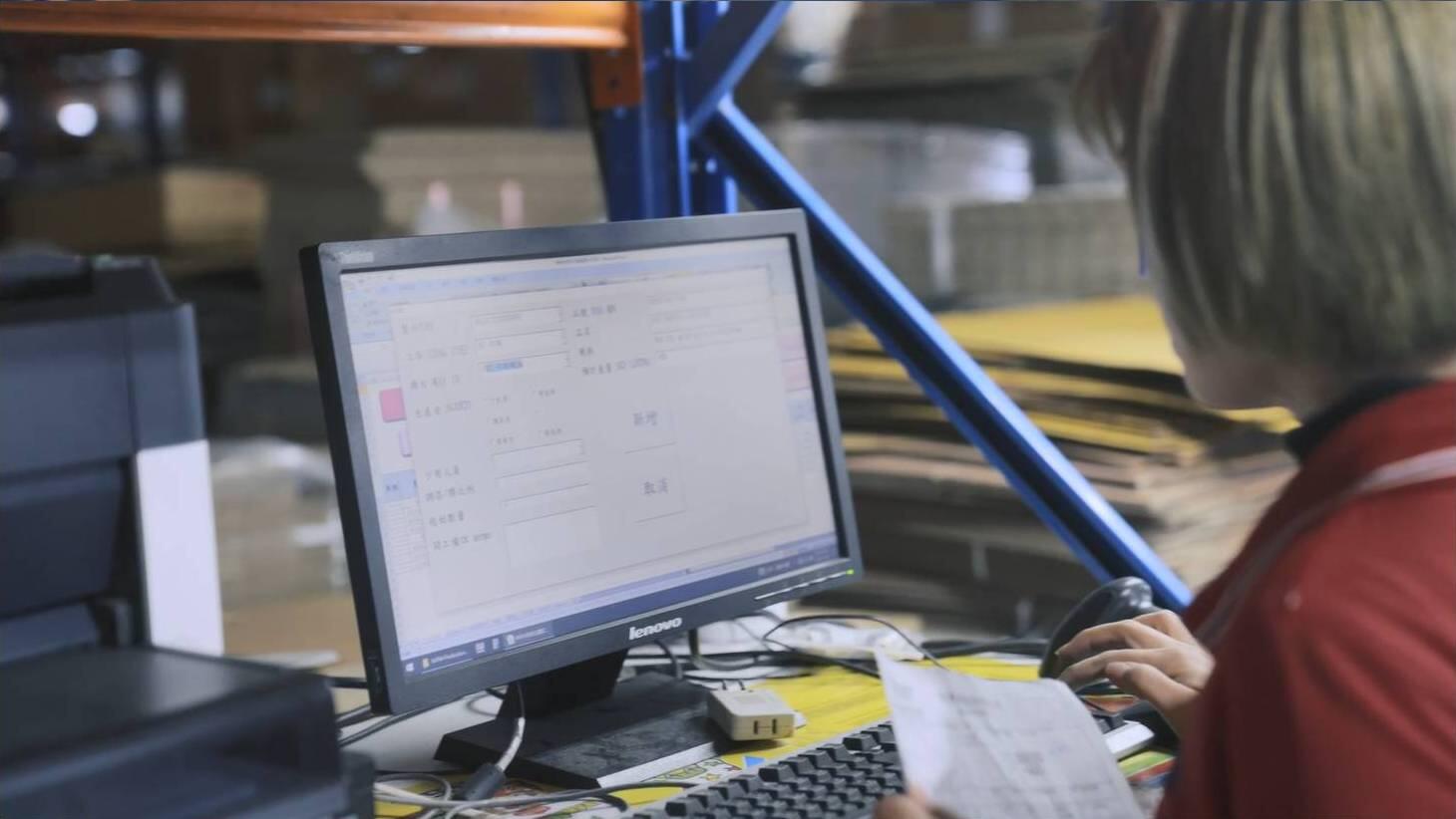 A person in a red shirt, likely a warehouse or logistics worker, sits at a desk in front of a Lenovo monitor, using a handheld barcode scanner to process documents or items.