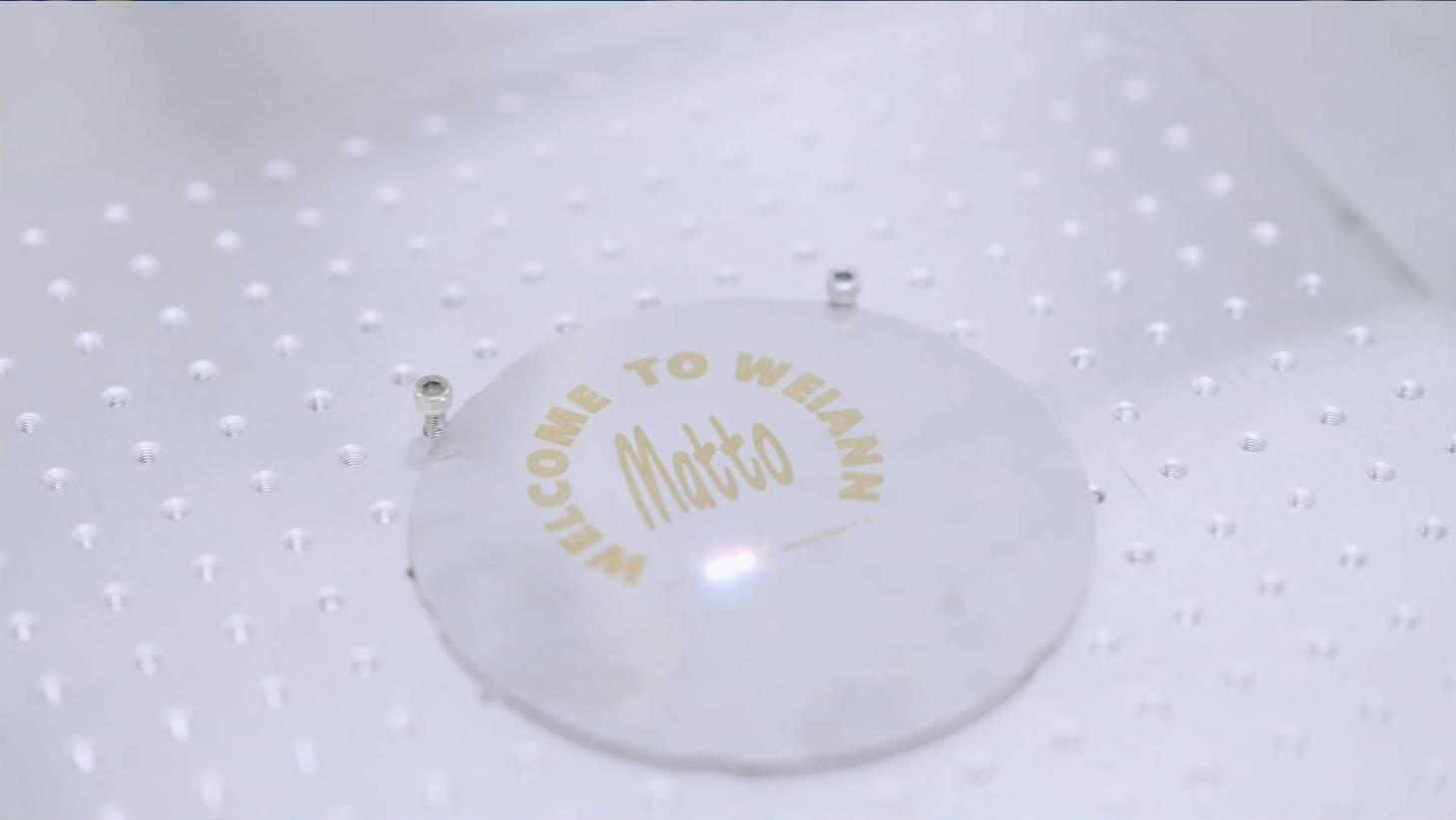 A close-up shot of a circular, clear object with "WELCOME TO WEIANN Matto" inscribed in gold lettering, placed on a translucent, textured surface with many small, raised dots. The sign is held in place by two small metallic fasteners.