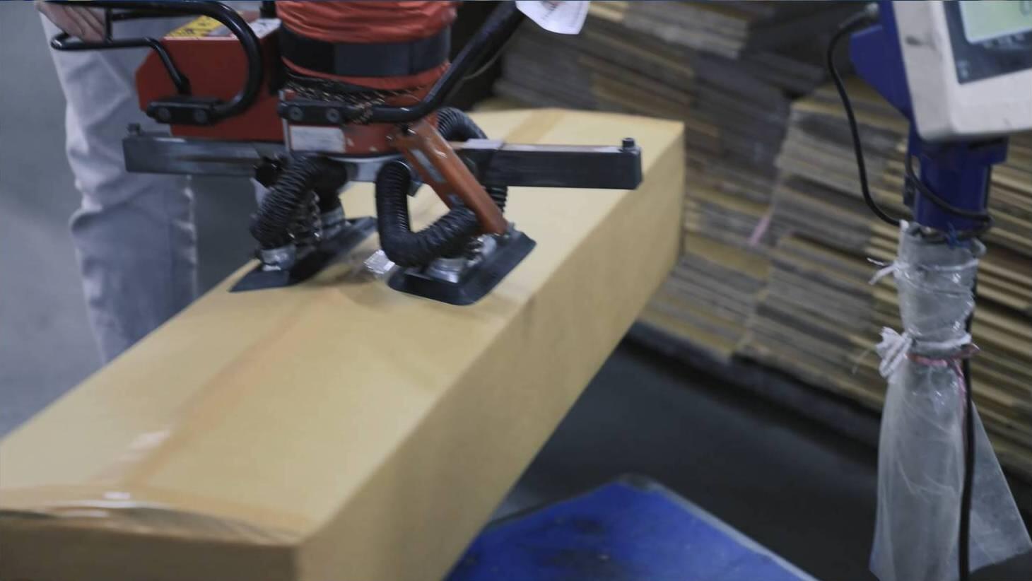 A powerful red and black industrial vacuum lifter, equipped with multiple suction cups, securely holds and lifts a large, unsealed cardboard box, likely in a warehouse or factory setting, demonstrating ergonomic material handling.
