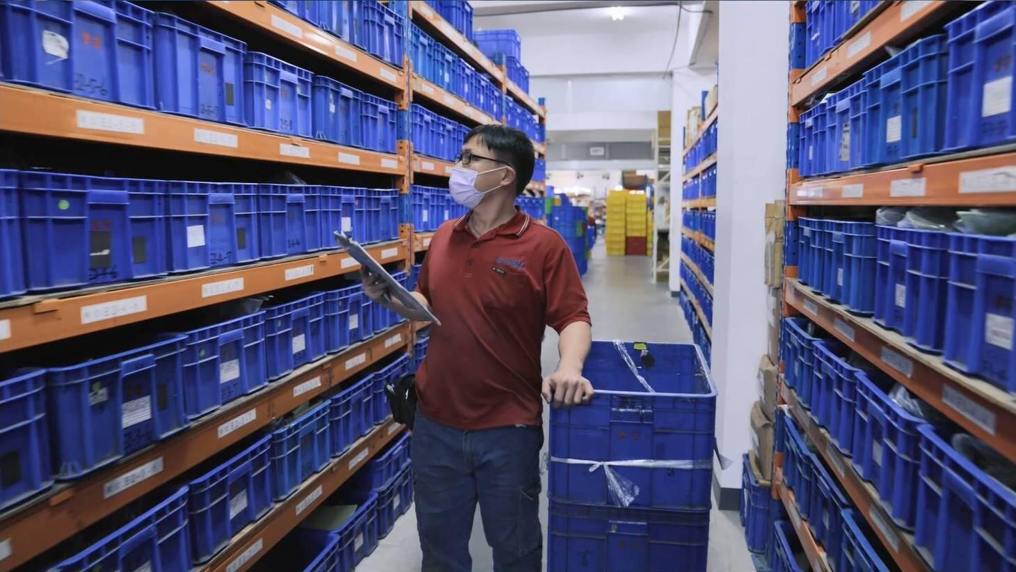 A man wearing a face mask and a red polo shirt stands in a narrow aisle of a warehouse, surrounded by shelves filled with blue storage bins. 