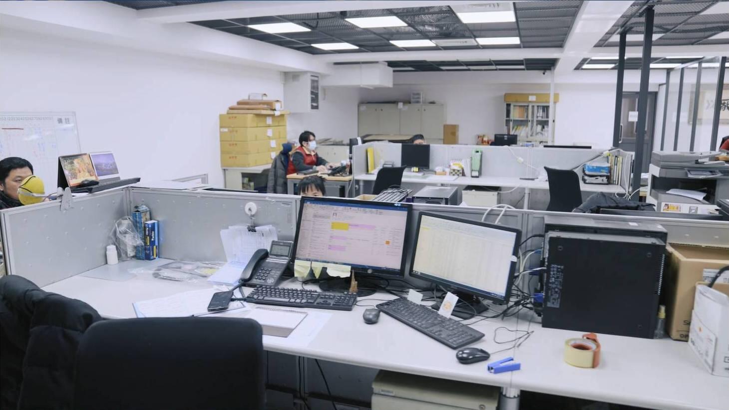 A wide-angle shot of a brightly lit, modern open-plan office space featuring several partitioned desks with computers, keyboards, and other office equipment.